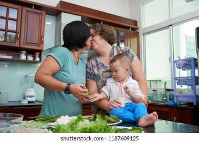 Lesbian Couple With Baby Boy Kissing When Cooking Dinner At Home