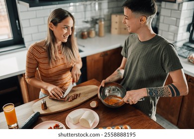 Lesbian couple happily preparing breakfast together in their modern kitchen, one whisking eggs while the other slices bacon, demonstrating a loving and domestic scene - Powered by Shutterstock