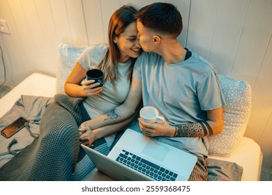 Lesbian couple holding hands, drinking coffee, and sharing intimate moments while using a laptop in bed, under a cozy blanket, enjoying their time together - Powered by Shutterstock