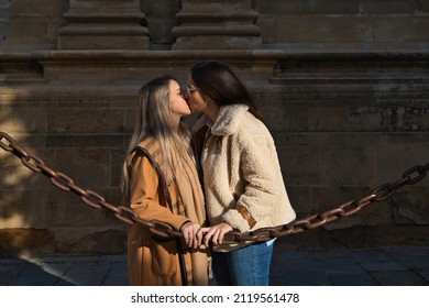 Lesbian Couple Leaning On A Chain Surrounding The Gothic Style Cathedral. The Women Are Kissing In Love And Passionately. Lesbian And Gay Marriage Concept.