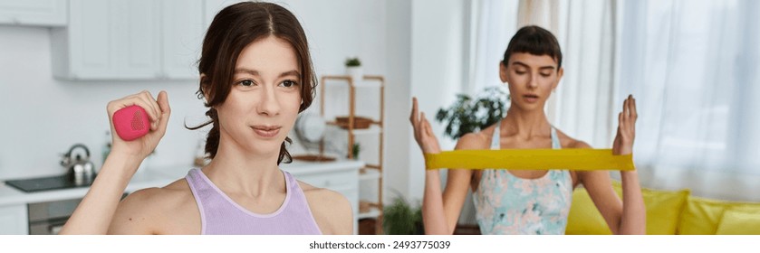 A lesbian couple works out in their apartment - one with a pink dumbbell and the other with a yellow resistance band - Powered by Shutterstock