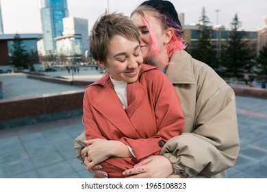 A lesbian girl with pink hair hugs her girlfriend with short hair while standing on a big city street. LGBT relationships. The concept of free love. High quality photo