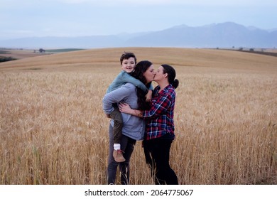 Lesbian Moms With A Young Boy. In A Wheat Field In The Country. The Boy Is Getting Piggyback From His Mother. The Moms Are Kissing.  