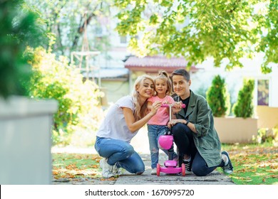 Lesbian Mothers With Young Daughter Walking With Scooter In The Park. Family Posing For A Portrait And Showing Cheerful Smiles.