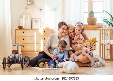 Lesbian Parents And Their Kids Playing With Toys In A Light Comfy Room. LGBT Family And Pride.