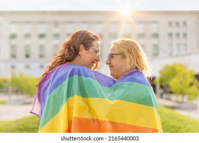 Lesbian women couple in love embracing each other, wrapped in a gay flag, in a park at sunset, with reflections of the sun. Concept of diversity, pride, love, equality.