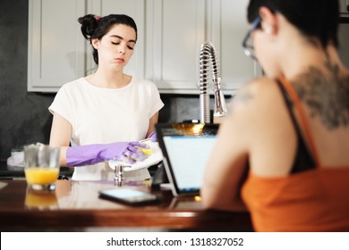 Lesbians At Home, Girl Washing Dishes In Kitchen, Partner Using Laptop Computer For Leisure. Young Hispanic Gay Woman Upset For Doing Home Chores Alone.