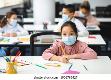 Lesson Concept. Portrait Of Little Asian Girl Wearing Single-use Protective Mask Sitting At Desk In Classroom Or Kindergarten, Writing Or Drawing In Notebook, Looking Away At Board. Return To School