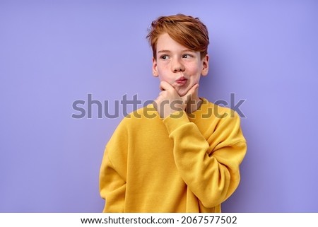Let me think. Caucasian pensive clever redhead schooler in yellow shirt with puzzled serious expression, child thinking doubting, making choice. indoor studio shot isolated on purple background