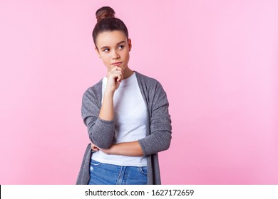 Let me think! Portrait of pensive cute brunette teenage girl with bun hairstyle in casual clothes holding hand on chin and thinking, pondering idea. indoor studio shot isolated on pink background