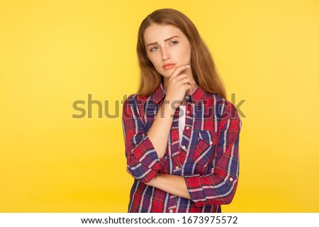 Let me think. Portrait of thoughtful ginger girl in checkered shirt standing with pensive look, pondering and musing, making plans, imagining in mind. indoor studio shot isolated on yellow background