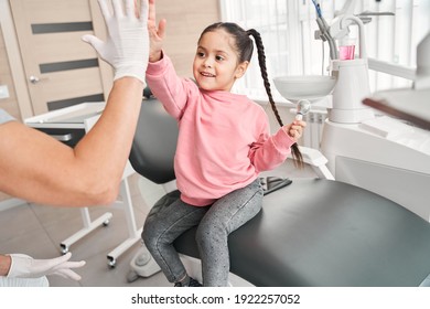 Let Us Be Friends Now. Emotional Cute Prominent Dentist Giving A High Five To Her Little Patient After Extracting Her Tooth. Happy Girl At The Dentist Chair. Stomatology For Kids Concept