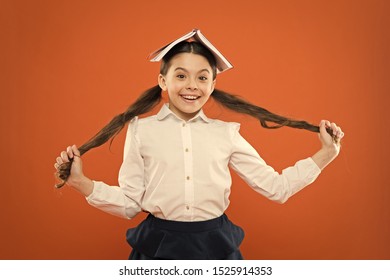 Let your hair speak for itself. Cute little girl holding long brunette hair on orange background. Small school child with book on head wearing hair in ponytails. Hair styling and haircare for school.