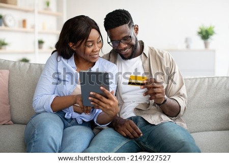 Let's Buy Something. Portrait of millennial black couple sitting on sofa at home and choosing what to order, excited woman pointing at digital tablet screen, guy in eyeglasses holding bankcard