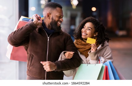 Let's Buy Something. Portrait of smiling black woman showing debit credit card to man, holding shopping bags, walking outside in the city near mall in the evening, blurred background