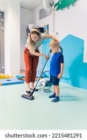 Let's Clean The Room, My Dear - Caucasian Boy In Blue Clothes Holding A Vacuum Cleaner And Cleaning The Floor With His Beautiful Teacher, Nursery Vertical Shot. High Quality Photo