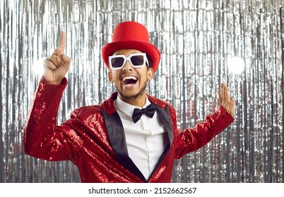 Lets get party. Positive funny young dark-skinned showman in shiny suit on silver foil background. Joyful crazy man in glasses, red hat and in suit with sequins raises his finger in sign of idea. - Shutterstock ID 2152662567