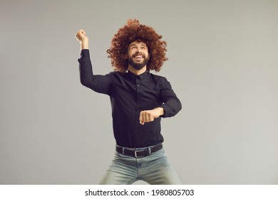 Let's Have A Party. Happy Carefree Man With Funny Crazy Hair Having Fun In Studio. Goofy Cheerful Young Guy Wearing Curly Brown Wig Dancing Gangnam Style Isolated On Gray Background