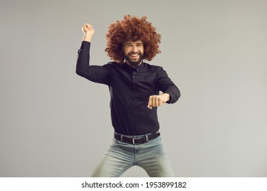 Let's Have A Party. Happy Young Man With Funny Crazy Hair Having Fun In Studio. Carefree Goofy Guy Wearing Curly Wig Smiling And Dancing Gangnam Style Isolated On Gray Background