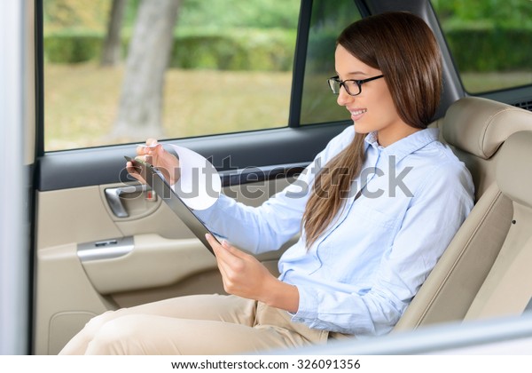 Lets plan the day. Pleasant upbeat positive young\
businesswoman holding folder and turning over the pages while\
sitting in the car
