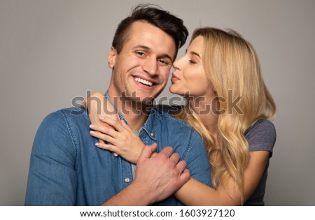 Let's smooch. Close up photo of an adorable woman, who is hugging her happy boyfriend in a denim jacket and trying to kiss him in the cheek.