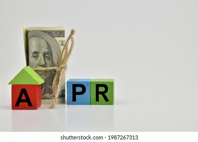 the letter APR printed on a wooden block, refers to the Annual Percentage Rate which is the interest rate charged in financing a housing loan. There is a dollar bill on the back of the wooden block.