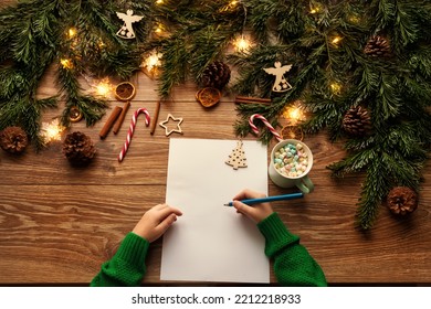 Letter to Santa. Christmas Child Hands Close up writing Xmas Gifts Shopping Wish List over brown Wooden Table. Wood Background with Fir Tree Branch decorated Christmas Lights and Toys