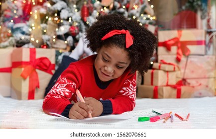 Letter To Santa. Cute Little Black Girl Lying On Floor And Writing Wish List Of Presents For Christmas In Decorated Room