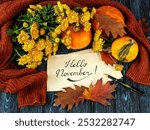 Lettering hello november on kraft paper, pen, pumpkins, orange sweater, autumn leaves, bouquet of yellow chrysanthemum flowers on wooden background