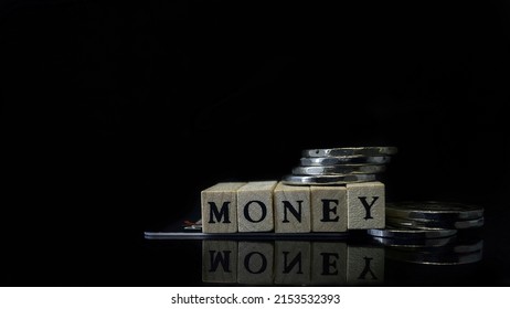 The Letters Or Spellings For The Word Money Is Isolated In Dark. Money Is Written On Wooden Cubes. A PAYONEER Bank Card And Coins Are Nearby. Selective Focus. Business Or Money Can't  Buy Happiness.