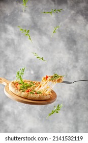 Levitation. Pizza with melted cheese, mozzarella, tomatoes, arugula and jalapeno pepper. Italian pizza on a wooden cutting board. Flying food. A slice of pizza on a shovel with stretching cheese. Grey