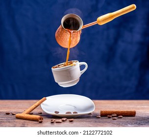 Levitación de verter café de una cezve en una taza con un salpicón. Palos de canela y granos de café sobre la mesa. Fondo azul, espacio de copia.