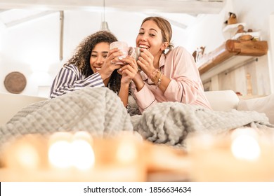 LGBT Female Couple Having Fun Under A Blanket On The Sofa - Cute Blonde Caucasian Woman Drink Cup Of Tea In Living Room Laughing With Her Black Hispanic Curly Girlfriend Just Woke Up In The Morning