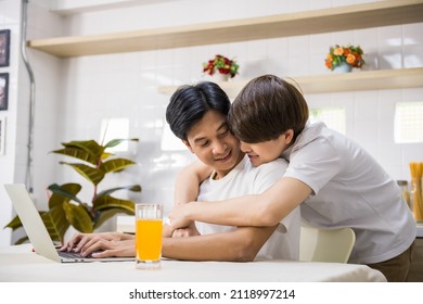 LGBT Man Work On Laptop While His Partner Hug Him From Behind At Living Room. Sweet Male Gay Couple Spend Time At Home. They Laugh And Look At Each Other. Same Sex Family.