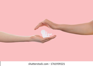 LGBT. Two Women's Hands Protect A Paper Baby. Copy Space. Pink Background. Concept Of Homosexual Families And Adoption