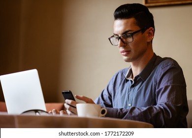 LGBTQ Community Lifestyle Concept. Young Homosexual Man Sits At The Table In Old-fashioned City Cafe. Handsome Gay Male Businessman Talks On A Phone While Having A Break At Lunch.