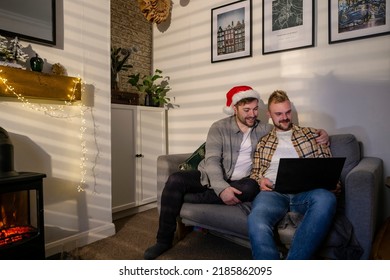 An LGBTQI Same Sex Male Couple Sitting On The Sofa In Their Living Room Relaxing, They Are Using A Laptop On A Video Call. One Man Is Wearing A Santa's Hat.