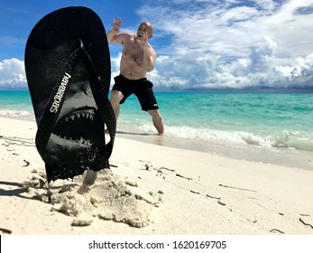  Lhaviyani Atoll, Maldives - October 12, 2019: Beach Slipper With Print. In The Background, A Man Pretends To Be Afraid Of A Shark. / Forced Perspective Photography. 