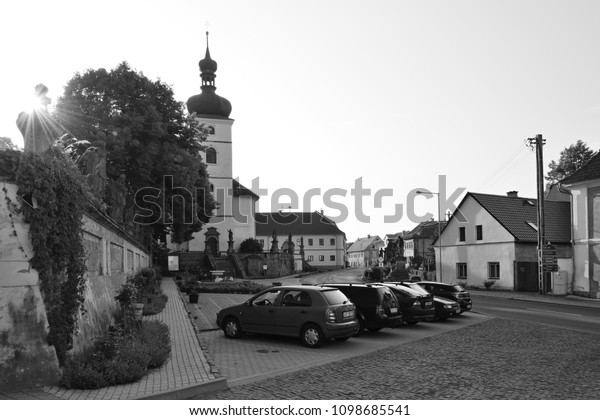 Libesice, Czech republic - May 19, 2018:\
parked cars between historical houses on little square with Kostel\
Nanebevzeti Panny Marie church on background during sunrise with\
black and white\
stylization