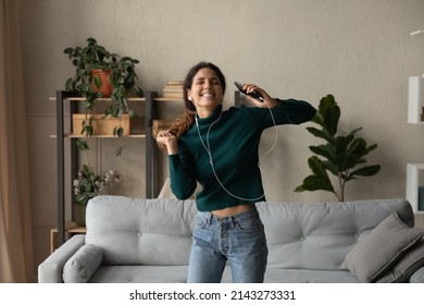 Life by music. Excited cheerful young latin female jump at living room by active rhythm from cellphone in wired headset. Cool energetic millennial woman move in crazy dance having fun relieving stress