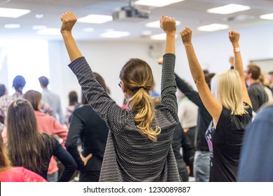 Life coaching symposium. Speaker giving interactive motivational speech at business workshop. Rear view of unrecognizable participants feeling empowered and motivated, hands raised high in air.