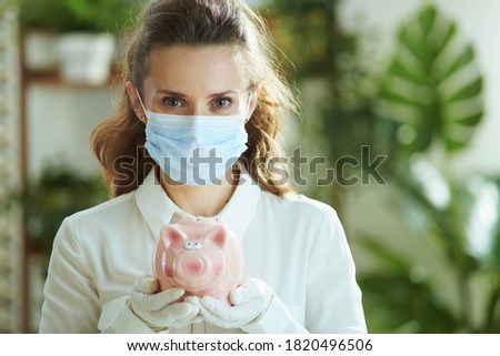 Life during coronavirus pandemic. Portrait of stylish middle age woman in white blouse with medical mask, piggy bank and gloves.