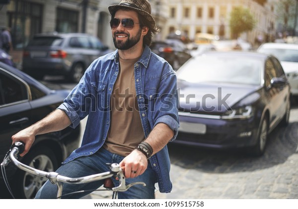 Life in movement. Portrait of cheerful man in\
headwear and sunglasses riding bike along the road. Cars on\
background. Copy space in right\
side