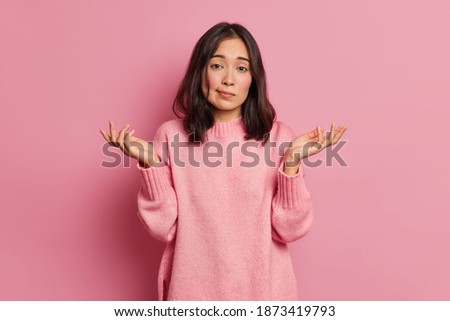 Life perception and hard decision concept. Puzzled clueless young Asian woman spreads palms and poses uncertian indoor expresses doubt wears knitted sweater isolated over pink studio background Сток-фото © 