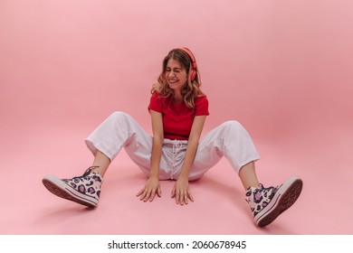 Life photograph happy young caucasian woman sitting on floor with legs spread wide against pink background. Cheerful blonde woman in casual clothes with headphones laughs hard. Playful mood concept