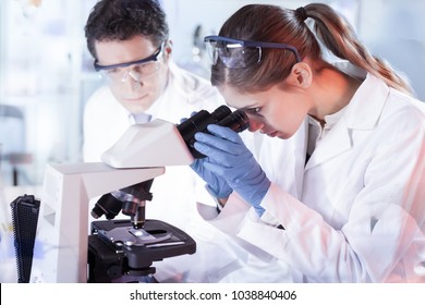 Life scientists researching in laboratory. Attractive female young scientist and her doctoral supervisor microscoping in their working environment. Healthcare and biotechnology.