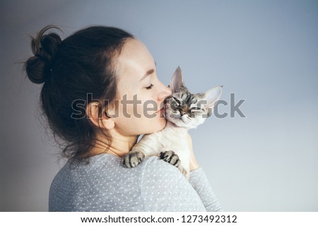 Life style photo of a casual dressed female who  is holding and petting and kissing cute Devon Rex cat. Kitten enjoys being in girls arms. Selective focus.