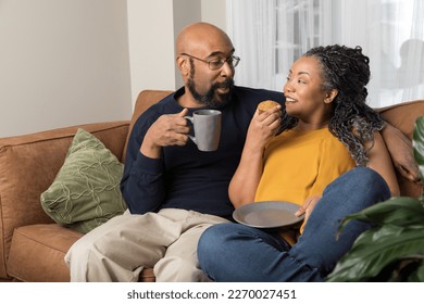 Life style portrait of a black married couple sitting on the couch and enjoying each other's company. The husband is drinking a coffee and the woman is eating a muffin.  - Powered by Shutterstock