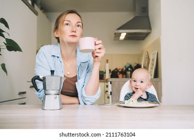 Life Of Working Mother With Little Baby. Young Tired Woman With Coffee And Baby Having Breakfast In Kitchen. Modern Freelancer Mom And Her Child After Sleepless Night. Work-life Or Work-family Balance