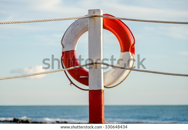 Lifebuoy white and red\
ropes hanging near the pier on the waterfront in Italy against the\
sky and the sea